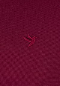 Bordeauxstof med en tekstureret overflade, prydet med et lille, broderet kolibri-logo i matchende farve.