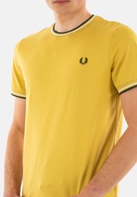 Camiseta amarilla de manga corta con escote redondo, que presenta acentos de rayas verdes y blancas en el cuello y las mangas, y un logo en el pecho.