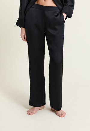 Kobieta ubrana w czarne satynowe spodnie o prostych nogawkach z kieszeniami, stojąca boso na tle jednolitego, jasnego tła.