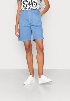 Personne portant un short bleu clair jusqu'aux genoux et des baskets blanches, avec une main dans la poche, devant un fond clair et uni.