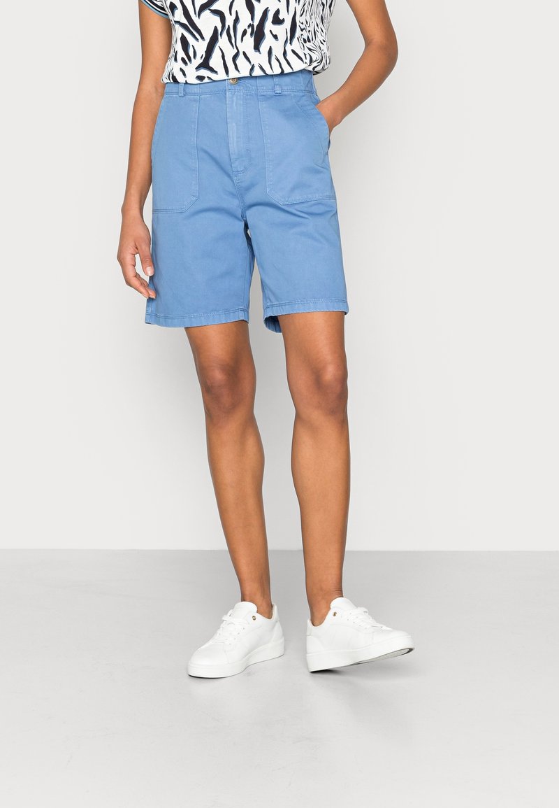 Persona che indossa pantaloni corti blu chiaro fino al ginocchio e sneakers bianche, con una mano in tasca, su uno sfondo chiaro e uniforme.