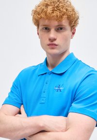 Νεαρός άνδρας με σγουρά κόκκινα μαλλιά, ντυμένος με φωτεινό μπλε πόλο μπλούζα Calvin Klein, στέκεται με τα χέρια σταυρωμένα μπροστά σε απλό φόντο.