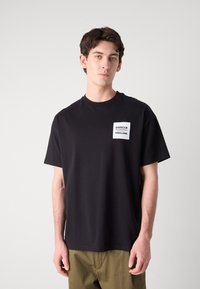 Zwarte katoenen t-shirt met ronde hals en korte mouwen, voorzien van een rechthoekig wit label met "BARBOUR INTERNATIONAL ENGLAND." Klassieke pasvorm.