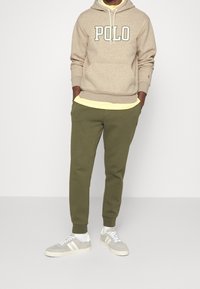 Béžová mikina s nápisem "POLO" v tmavě zelené barvě, navlečená přes žluté tričko. Olivově zelené tepláky a světle šedé tenisky s bílými pruhy.