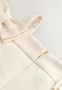Kreemjas kleidis on tekstuuriga satiinviimistlus. Sellel on paela detail ja kuldne tõmblukk külje peal. Lihtne, elegantne disain ning reguleeritavad rihmad.