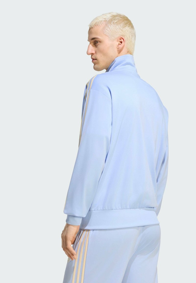 Blonde man wearing a light blue tracksuit with beige stripes, standing sideways and looking to the left against a plain background.