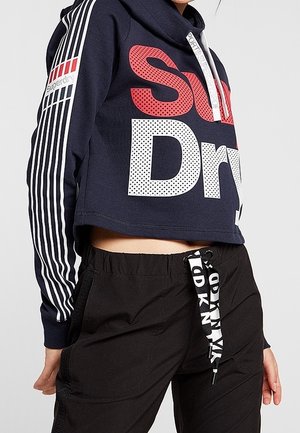 Osoba ubrana w granatową, krótką bluzę z kapturem z logo "Superdry" i prążkowanymi rękawami, zestawioną z czarnymi spodniami dresowymi z troczkiem z logo DKNY.