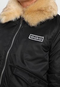 Černá bomber bunda s límcem z umělé kožešiny, se zipem a nášivkou s čísly na levé straně. Hladká textura, minimalistický design.