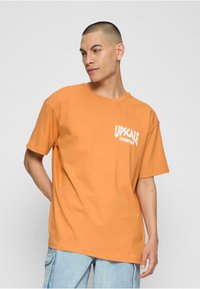 Oranje katoenen t-shirt met een ronde hals en korte mouwen. "UPSCALE STUDIOS" logo in het wit gedrukt op de linkerkant van de borst.