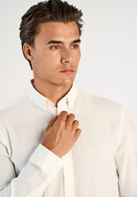 Camicia bianca con bottoni in tessuto strutturato, taschino sul petto singolo, colletto con bottoni e maniche lunghe. Modello che aggiusta il colletto con una mano.