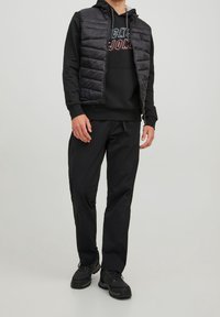 Gilet puffer nero sopra un hoodie nero con scritte, abbinato a pantaloni jogger neri e sneakers nere. Design semplice e funzionale.