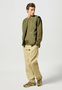 Pikowany zielony kamizelka na oliwkowym swetrze, w zestawieniu z beżowymi spodniami cargo oraz butami sportowymi w brązowo-szarym i zielonym wzorze.