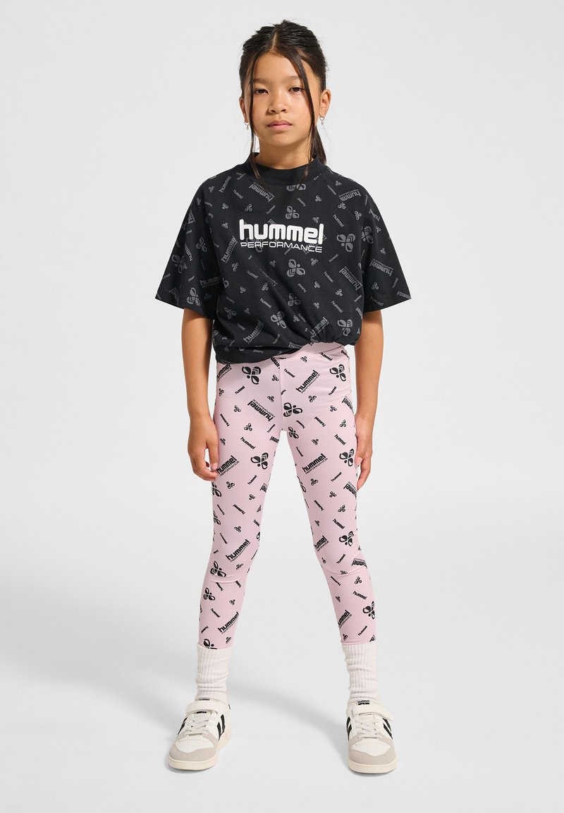 Czarny krótki t-shirt z powtarzającym się wzorem logo. Jasnoróżowe legginsy z pasującymi nadrukami logo. Białe trampki i teksturowane skarpetki.