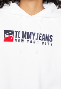 Sudadera blanca con el logo de Tommy Jeans de Nueva York bordado en navy y rojo en el pecho, llevada por una persona con cabello rizado y oscuro.