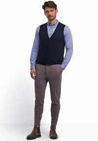 Gilet a maglia blu navy indossato sopra una camicia azzurro chiaro, abbinato a pantaloni grigi a sigaretta e stivaletti marroni. Tessuto liscio con una texture sottile.