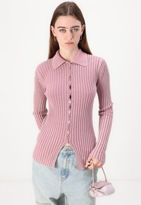 Top de malha canelada rosa com colarinho, apresentando uma frente com botões e bainha com racha. A textura é suave, com um design ajustado e mangas longas.