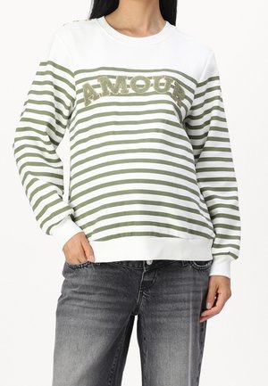 Femme portant un t-shirt à manches longues rayé vert et blanc avec le texte « AMOUR » sur la poitrine et un jean noir délavé, main dans la poche.