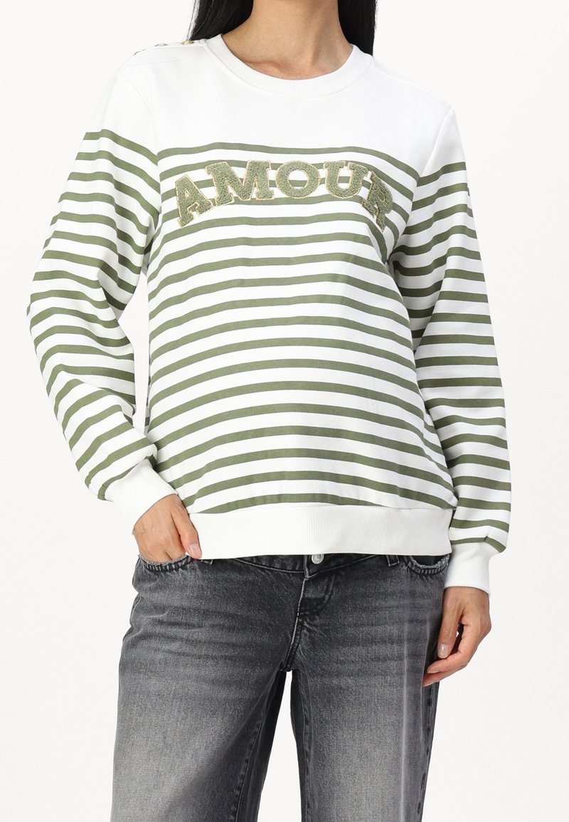 Femme portant un t-shirt à manches longues rayé vert et blanc avec le texte « AMOUR » sur la poitrine et un jean noir délavé, main dans la poche.