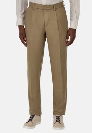 Hombre vistiendo pantalones marrón claro a medida combinados con zapatillas beige y una camisa blanca de manga larga, de pie con los brazos relajados.