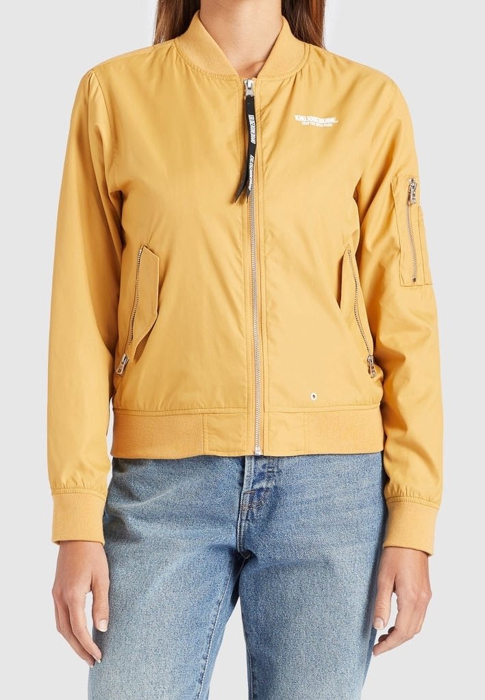 Veste bombardier jaune avec fermeture éclair à l'avant, col côtelé, poches latérales et poche à fermeture éclair sur la manche. Fabriquée en tissu lisse et léger.