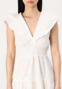 Balta suknelė su giliu V formos iškirpimu, raukšlėtais petnešėlėmis ir sluoksniuota sijona, pasižyminti švelnia, lengva medžiaga ir laisvu kirpimu.