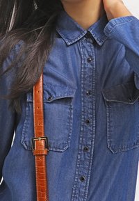 Denimová košile v tmavě modré barvě s límcem, dvěma náprsními kapsami a předním zapínáním na knoflíky. Hnědý texturovaný pás je částečně viditelný.