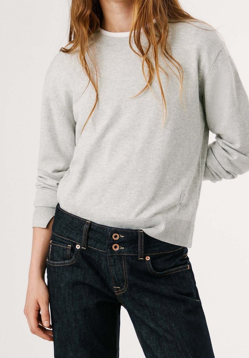 Femme portant un pull gris clair à manches longues, légèrement rentré dans un jean taille haute foncé avec des boutons doubles.