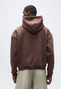 Άνδρας με καφέ φούτερ με κουκούλα κατεβασμένη, που στέκεται με την πλάτη προς τα εμπρός, συνδυασμένος με ανοιχτόχρωμο μπεζ παντελόνι, πάνω σε απλό φόντο.