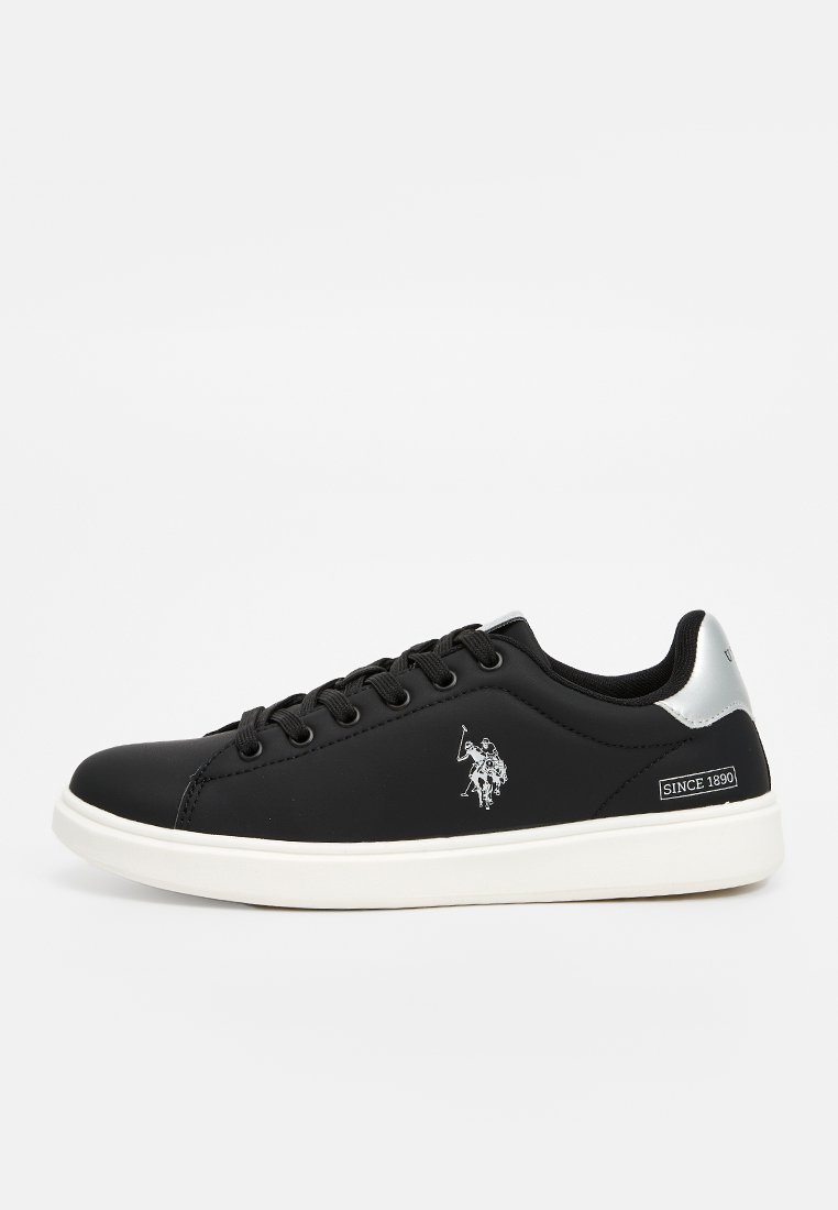 Zapatilla sintética negra con suela de goma blanca, con detalles plateados en el talón y un logo blanco en el lado exterior.