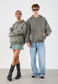 Szürke, cipzáros kapucnis felsők, oversized fazonban, kenguru zsebekkel. A modellen farmer szoknya és fekete térd feletti csizmák vannak; a másodikon világos farmer és sportcipők.