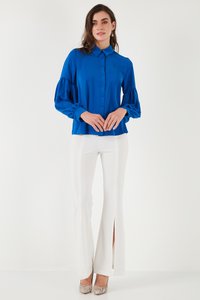 Žena koja nosi svijetloplavu bluzu dugih rukava s gumbima i bijele hlače visokog struka s prednjim prorezima, stoji ispred bijele pozadine.