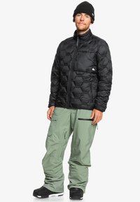 Zwarte pufferjas met hexagonaal patroon, voorzien van een rits aan de voorkant en zijzakken, gecombineerd met lichtgroene sneeuwbroeken en zwarte laarzen.