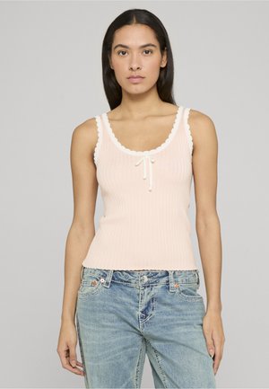 Mujer con una camiseta sin mangas acanalada de color rosa pálido con bordes en forma de onda y un pequeño lazo, combinada con unos vaqueros azul claro, posando frente a un fondo gris.