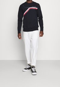 Navy sweatshirt with geometric color block detailing in red and white. Paired with white pants and black sneakers featuring white laces.