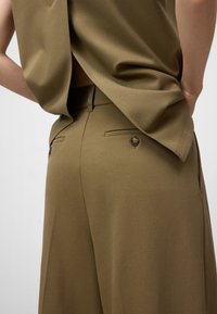 Olivgröna skräddarsydda byxor med knappade bakfickor, kombinerat med en matchande lös topp, händerna vilande bakom midjan.