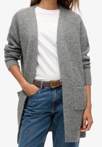 Gray knitted cardigan with open front, rolled sleeves, and two front pockets, worn over a white shirt and blue jeans.