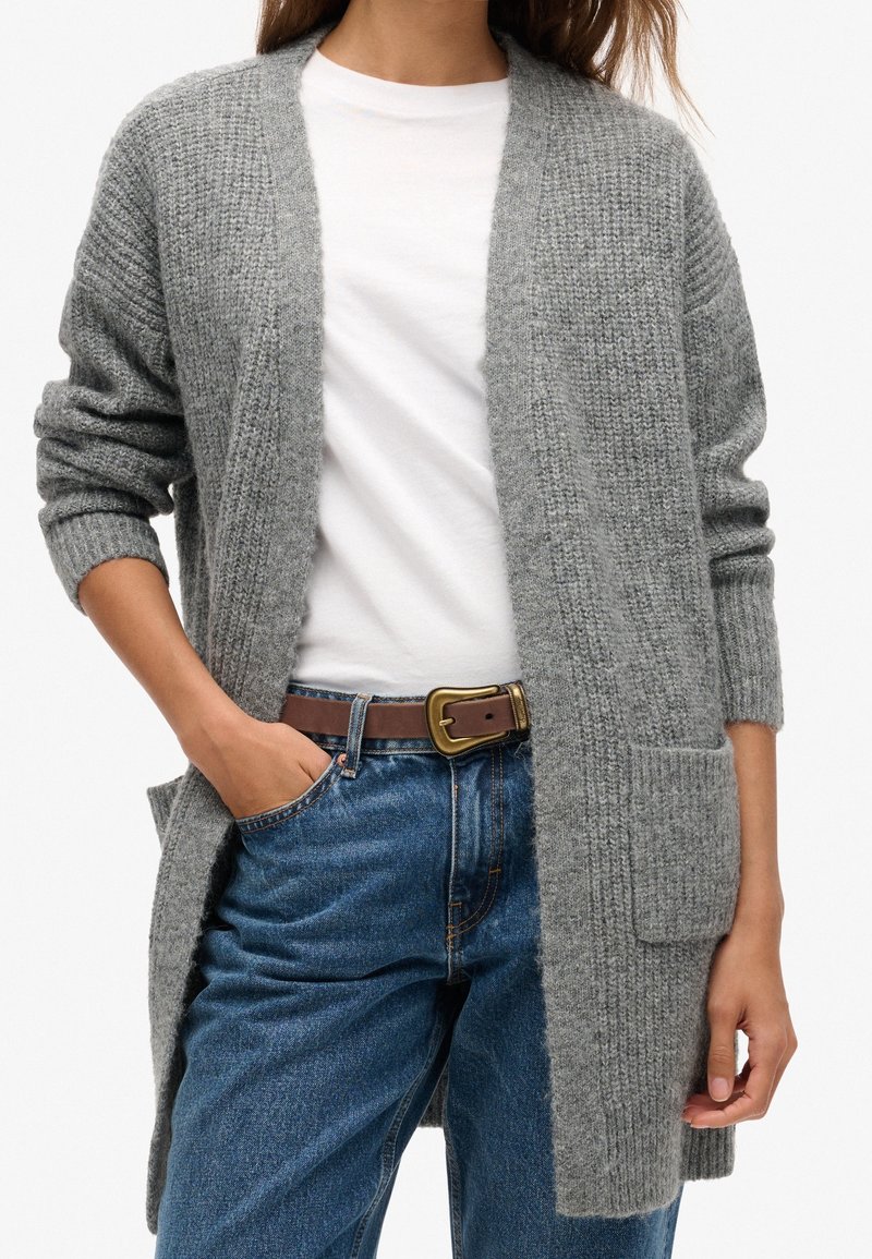 Gray knitted cardigan with open front, rolled sleeves, and two front pockets, worn over a white shirt and blue jeans.