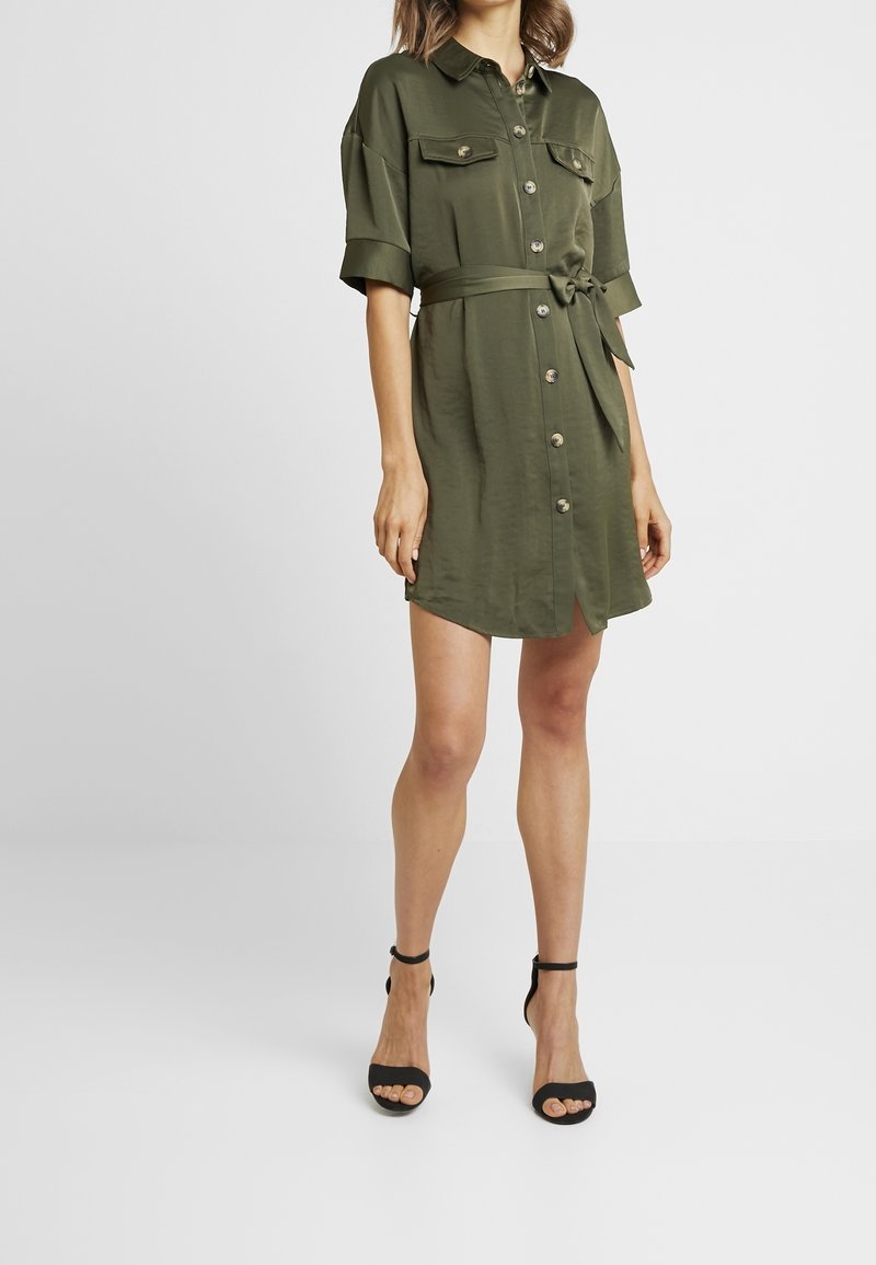 Femme portant une robe-chemise à manches courtes vert olive avec ceinture nouée à la taille et sandales à talons hauts noires avec bride à la cheville.
