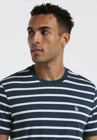 Jeune homme avec des cheveux bouclés courts portant un t-shirt rayé horizontal bleu marine et blanc avec un petit logo de pingouin sur la poitrine.