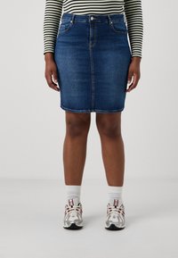 Saia de ganga, azul-escuro, até ao joelho, com cinco bolsos, bainha costurada, usada com meias brancas e ténis de cores mistas.