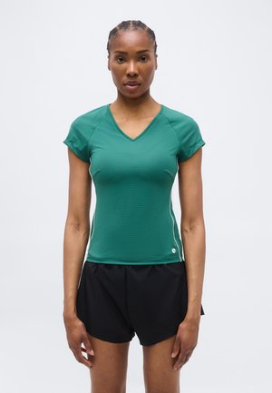 Woman standing facing forward wearing a green fitted V-neck short-sleeve top and black athletic shorts against a plain background.