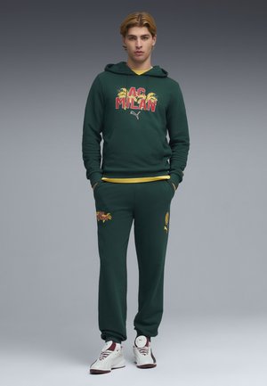 Jonge man met handen in zijn zakken, gekleed in een donkergroene AC Milan Puma hoodie en bijpassende joggingbroek, met witte sneakers.
