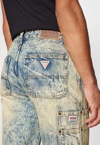 Lyseblå denimjeans med en falmet tekstur, som har to baklommer og en rød trekantlogo. Brun lærmerkelapp i midjen.