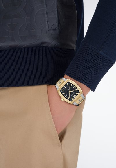 Granatowy sweter z prążkowanymi mankietami; granatowa skórzana wstawka z wzorem. Złoto-srebrny zegarek z czarną tarczą i prostokątną kopertą na nadgarstku.