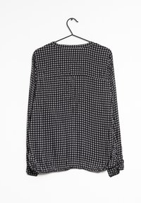 Blouse à manches longues à motifs noir et blanc avec un col rond, tissu léger et ourlet arrondi, suspendue à un crochet noir.