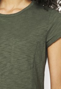 T-shirt vert à manches courtes en tissu texturé, avec une petite poche sur la poitrine gauche et un col rond.
