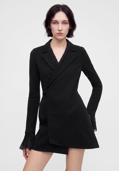 Jeune personne aux cheveux courts et foncés portant une robe blazer asymétrique noire avec des poignets plissés transparents, debout devant un fond uni.