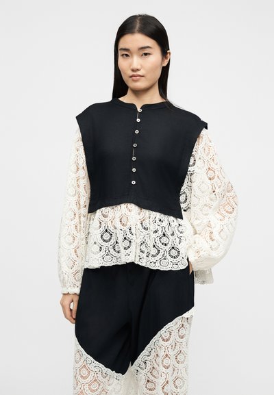 Γυναίκα με μαύρο κοντό αμάνικο τοπ πάνω από λευκό δαντελένιο πουκάμισο με φουσκωτά μανίκια και ταιριαστό μαύρο παντελόνι με λευκές δαντελένιες επιφάνειες.