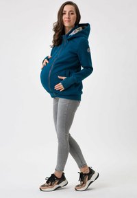 Felpa con zip di colore teal con una fodera interna a motivo, vestibilità comoda, tasche laterali e bordi a costine, indossata con jeans skinny grigi e sneaker multicolori.
