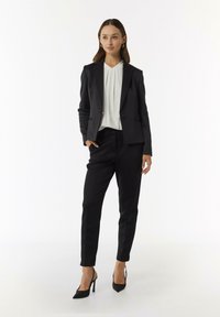 Schwarze taillierte Blazer über einer weißen Bluse, kombiniert mit maßgeschneiderten schwarzen Hosen und schwarzen hohen Schuhen. Glattes Material mit strukturiertem Design.